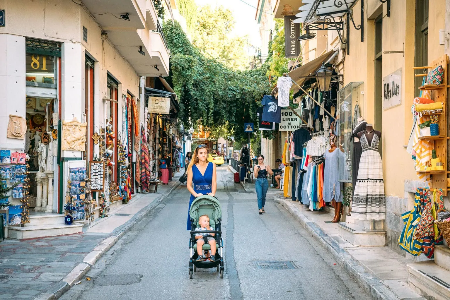 Mom pushing a stroller down a street with local shops in Greece, an example of exploring with a toddler.
